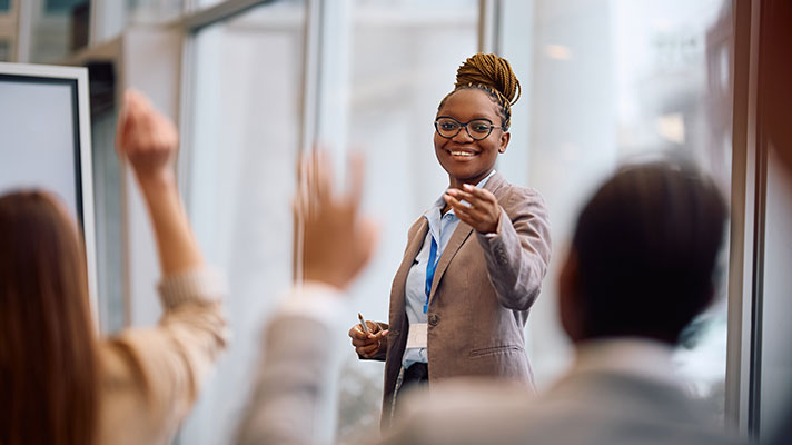 A black female stands in front of a group of people pointing to someone with their hand up