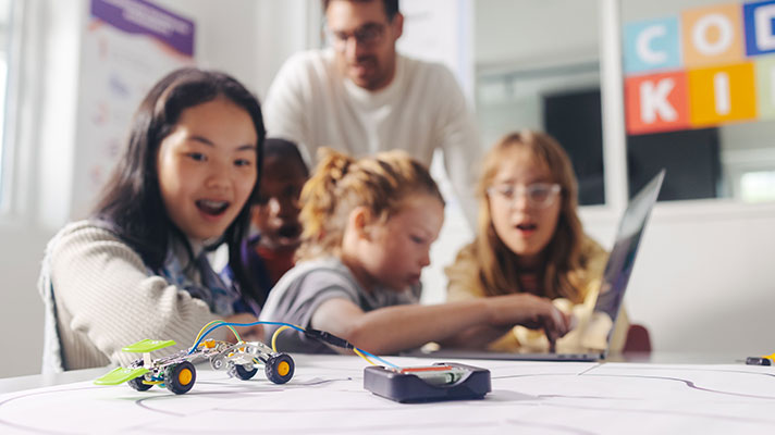 A robotic vehicle is shown in the foreground, three students and teacher are shown in the background. One student works on a laptop