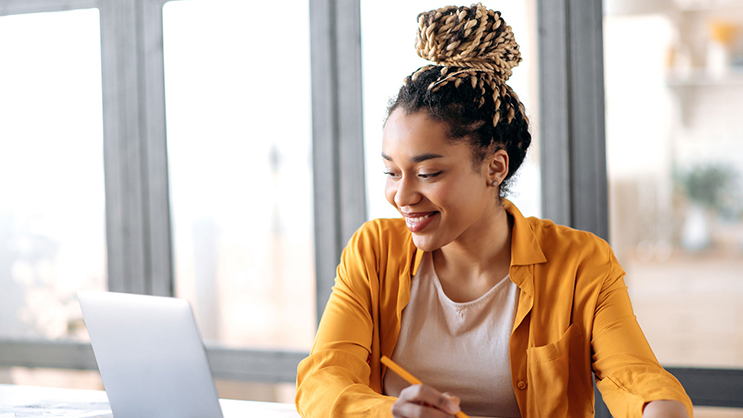A young black woman smiles at her laptop screen while writing in a notebook. She is wearing a white undershirt and an unbuttoned orange dress shirt.