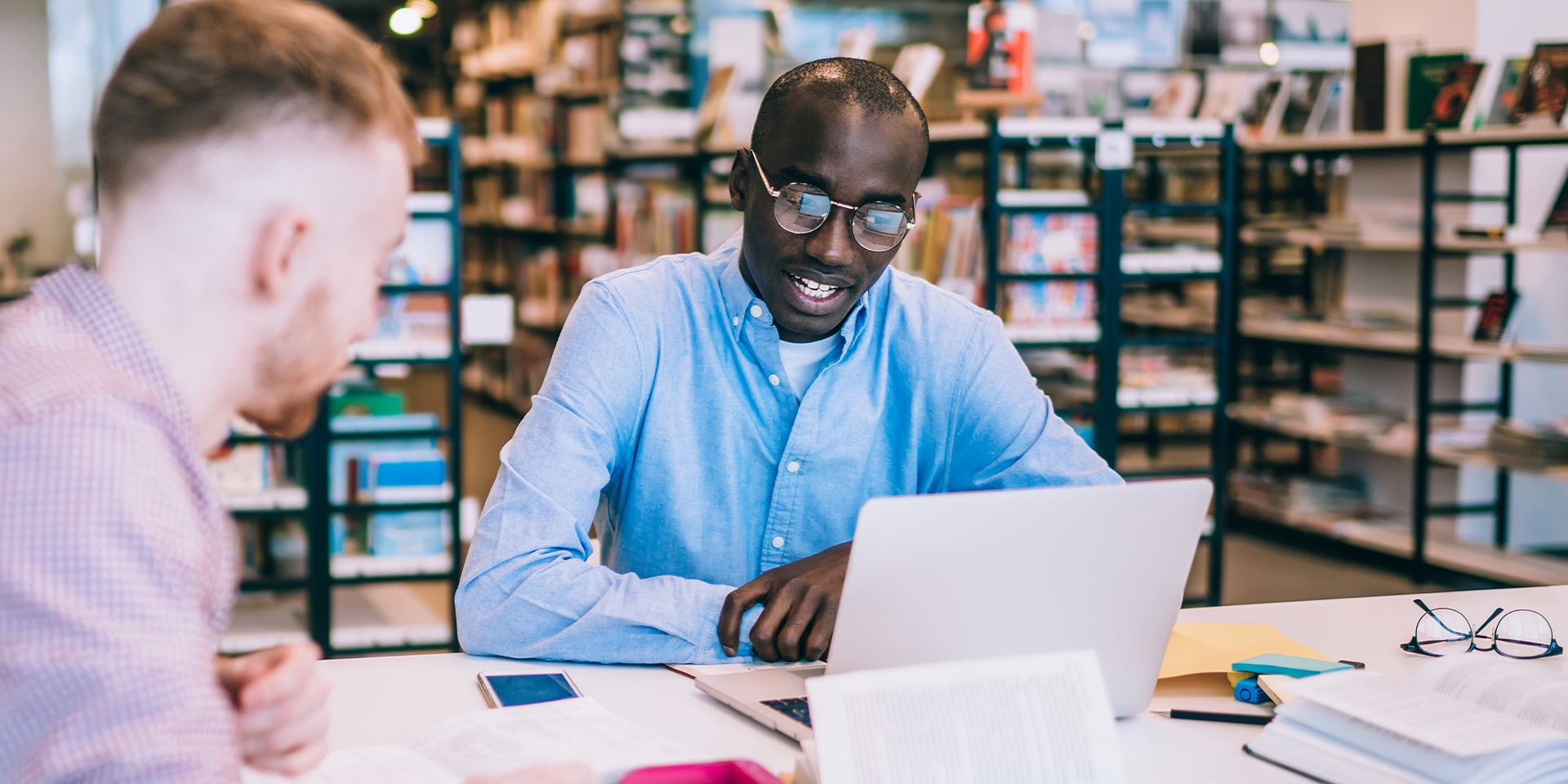 Two men dressed in business casual attire chatting with each other. They are seated at a table full of laptops, textbooks, folders, and other office supplies.