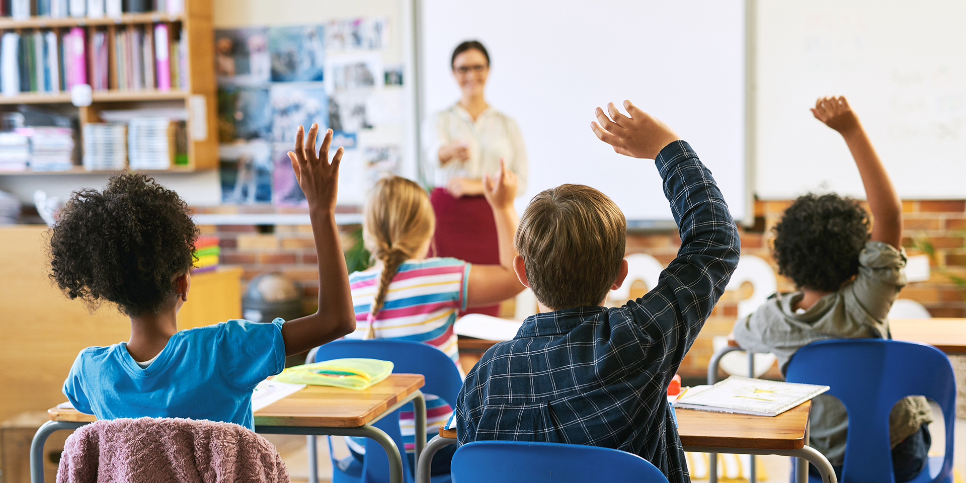 Students raise their hands while a teacher stands at the front of the classroom in the background.