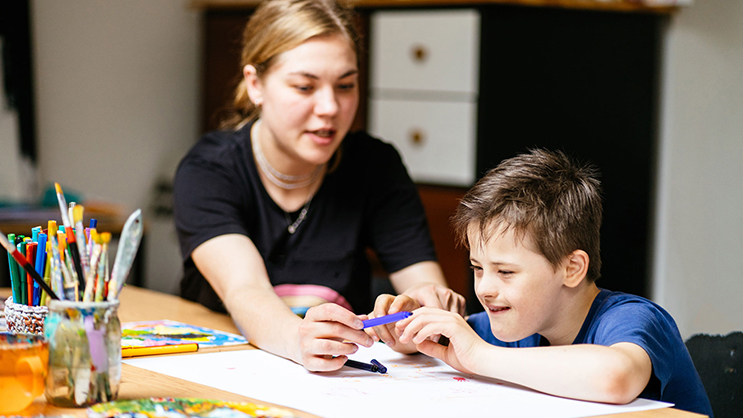 A young teacher helps a special needs student. The teacher is a white woman wearing a black shirt and necklaces. The student is wearing a royal blue t-shirt and looking at a purple marker. 