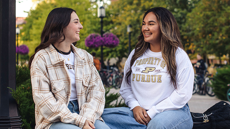 Two female Purdue students seated outdoors. They are both wearing jeans and Purdue attire. 