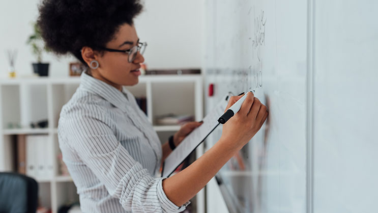 A teacher writing on a whiteboard with a black dry erase marker. She is wearing glasses and a striped white dress shirt.