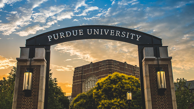 A dimly illuminated silhouette of the Purdue University Arch in front of a cloudy sunset.