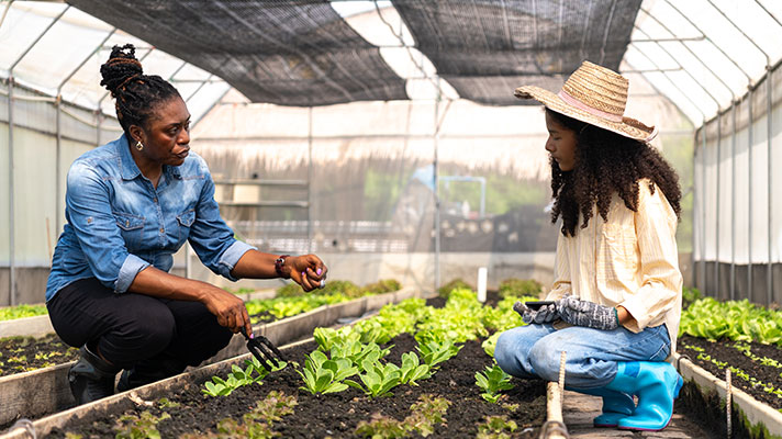 A teacher works with a student and green plants in a greenhouse