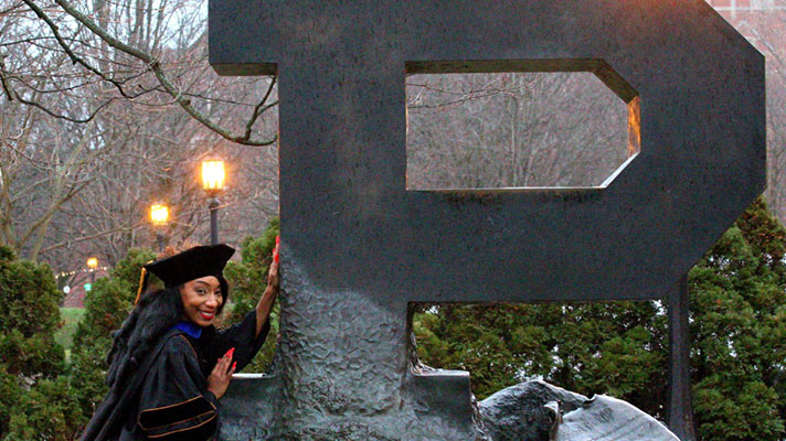 A PhD student stands in front of the large Purdue 
