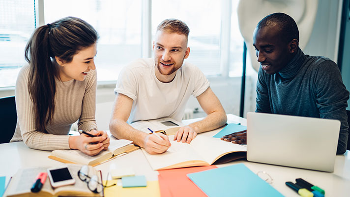 Three students work on a project. One student writes in a notebook and another student works on a laptop