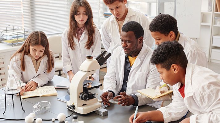 A teacher in a white lab coat works with students in white lab coats around a microscope