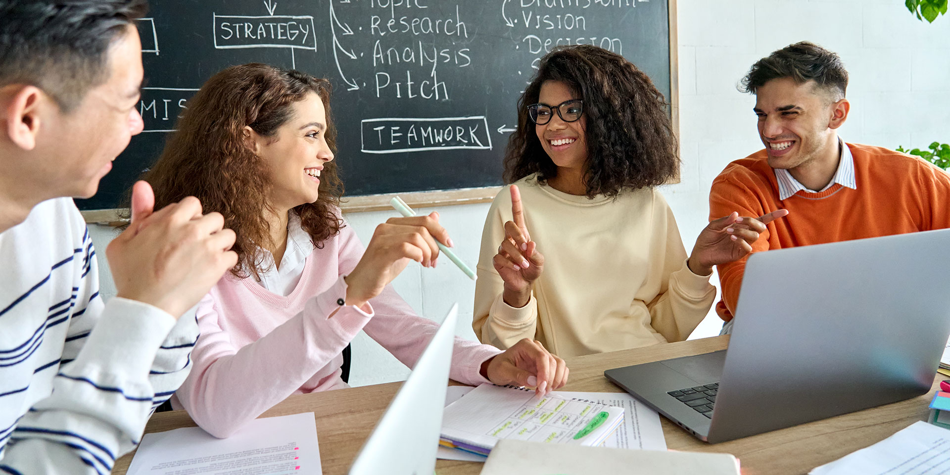A group of students work on a group project. Behind them is a chalkboard and in front of them on a table is a laptop and notebook.