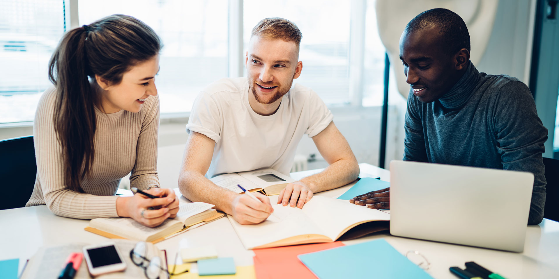 Three students work on a project. One student writes in a notebook and another student works on a laptop.