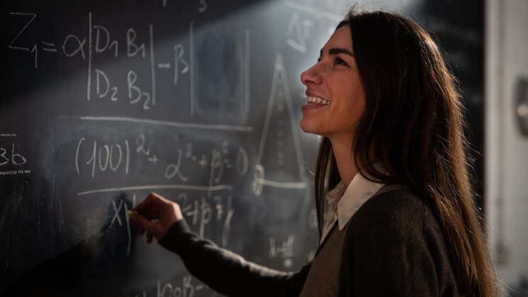 A teacher writes on a chalkboard. She has long brown hair and is wearing a white shirt, beige vest, and brown cardigan.