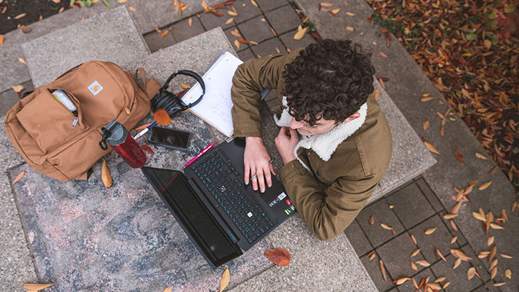 An aerial view of a Purdue student seated outside. On a table in front of him are his backpack, water bottle, headphones, cell phone, notebook, and laptop.