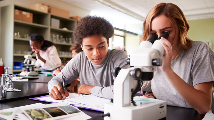 Two students sit at a table, one takes notes, the other looks through a microscope.