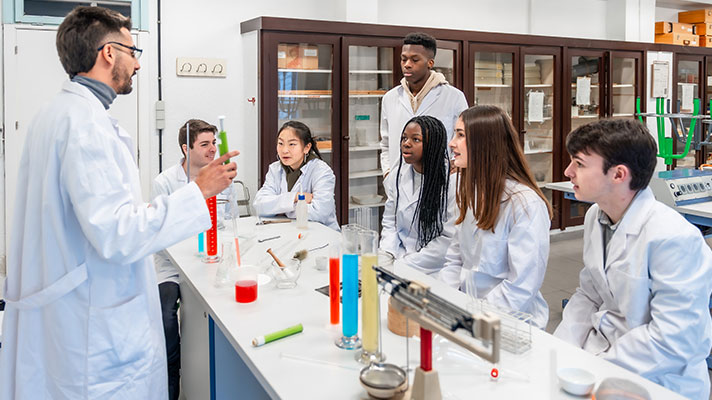 A teacher in a white lab coat explains a chemistry lab experiment to a group of students in white lab coats