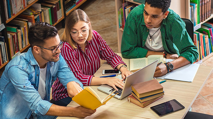 Three students work with books and laptop in a library