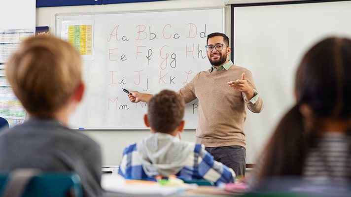 A male teacher points to a letter in the alphabet on the whiteboard.