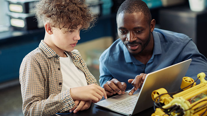A black male teacher works with a student on a laptop.