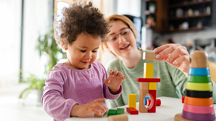 A child plays with blocks while a woman helps in the background
