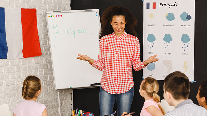 A teacher stands in front a white board with a French lesson. A France flag is shown in the background.