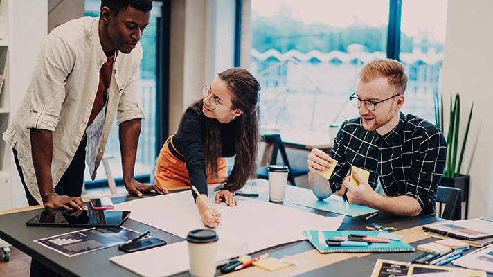 Three students work on a project on a large table, coffee cups, papers and sticky notes are strewn across the table