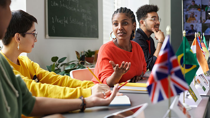A student talks to other students during a project, different world flags are shown in the foreground