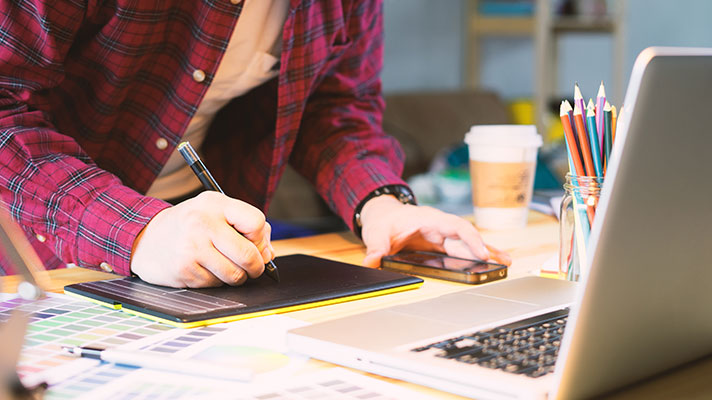 A person works on a tablet with color swatches and colored pencils shown in the foreground.