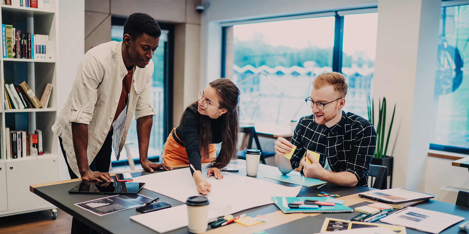 Three students work on a project on a large table, coffee cups, papers and sticky notes are strewn across the table