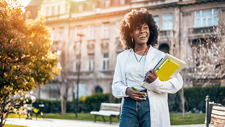 A young college student outside holding a notebook and textbook in her arms and smiling.