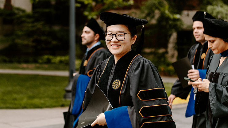 A graduate student with glasses wearing her commencement attire. She is outside with other graduate students in the background.