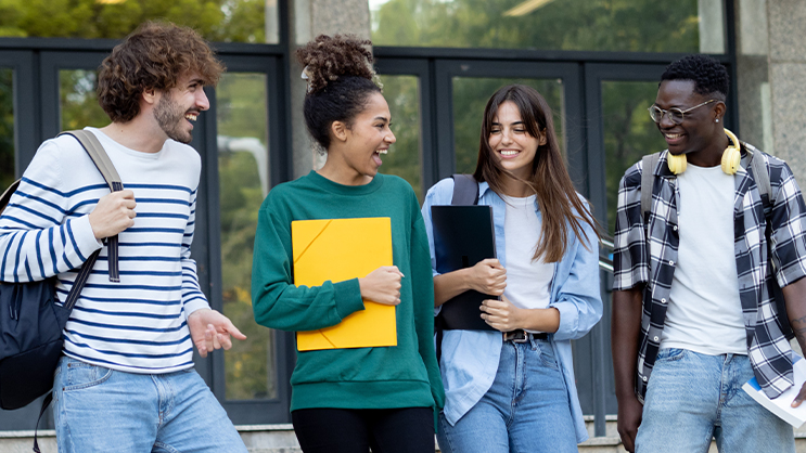 A diverse group of four college students laughing outside of a building. They are holding notebooks and wearing backpacks.