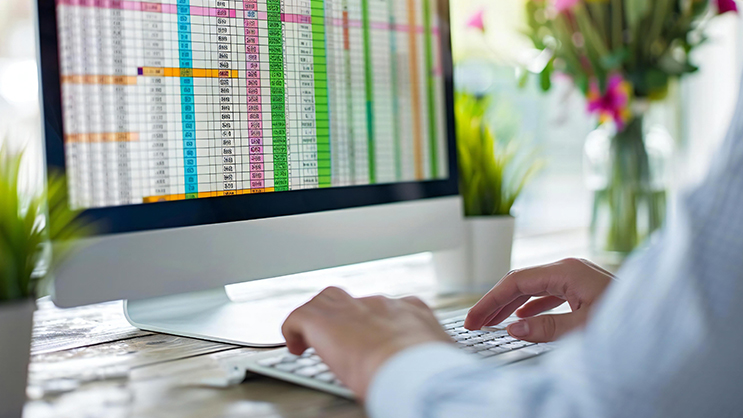 A businessman typing on a keyboard. In front of him is a computer monitor with a color coded spreadsheet visible.