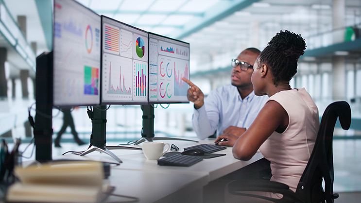 Two young professionals viewing charts and graphs on a set of three computer monitors.