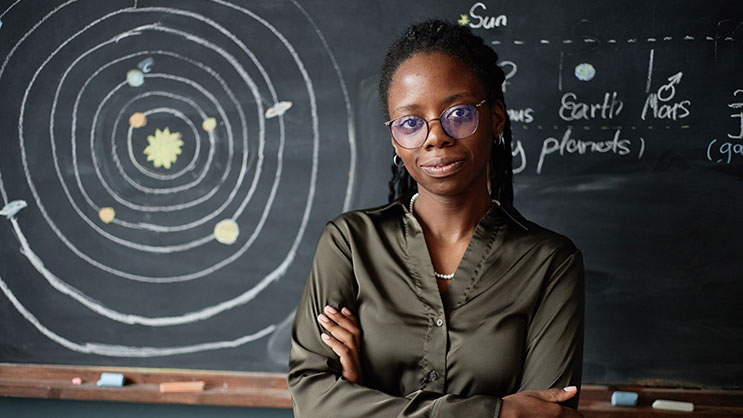 A black female teacher stands in front of a drawing of the solar system on a chalkboard