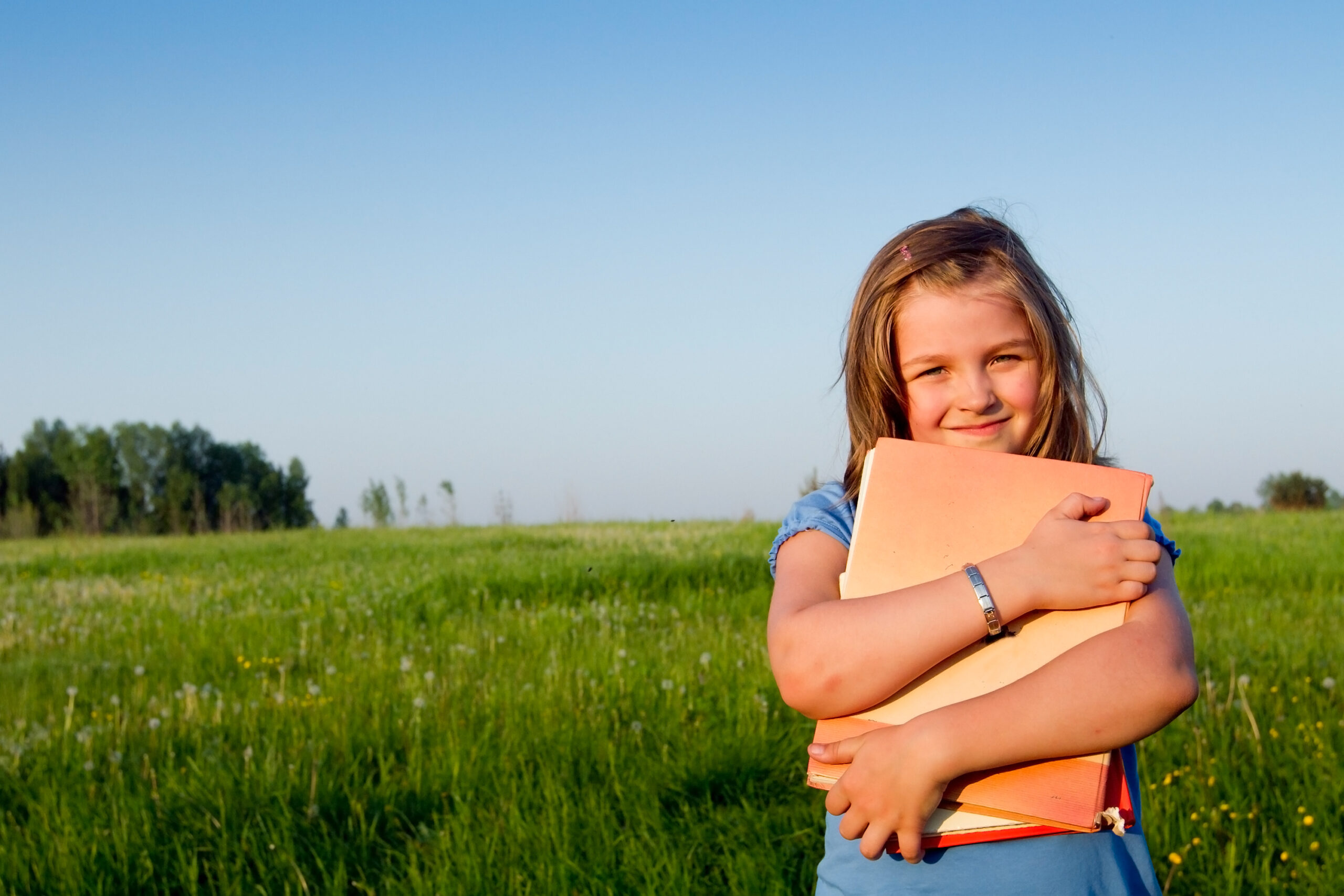 A young white girl stands in a green field of grass holding a notebook