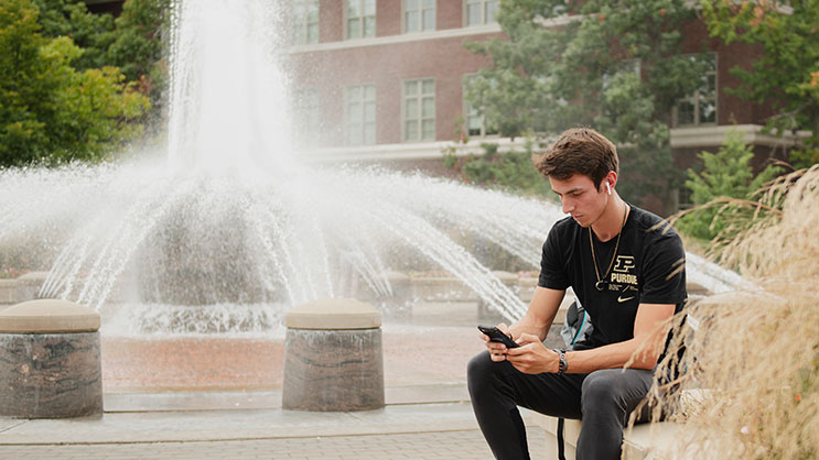 A white male Purdue University student sits in front of Loeb Fountain looking at his phone with an AirPod in his ear
