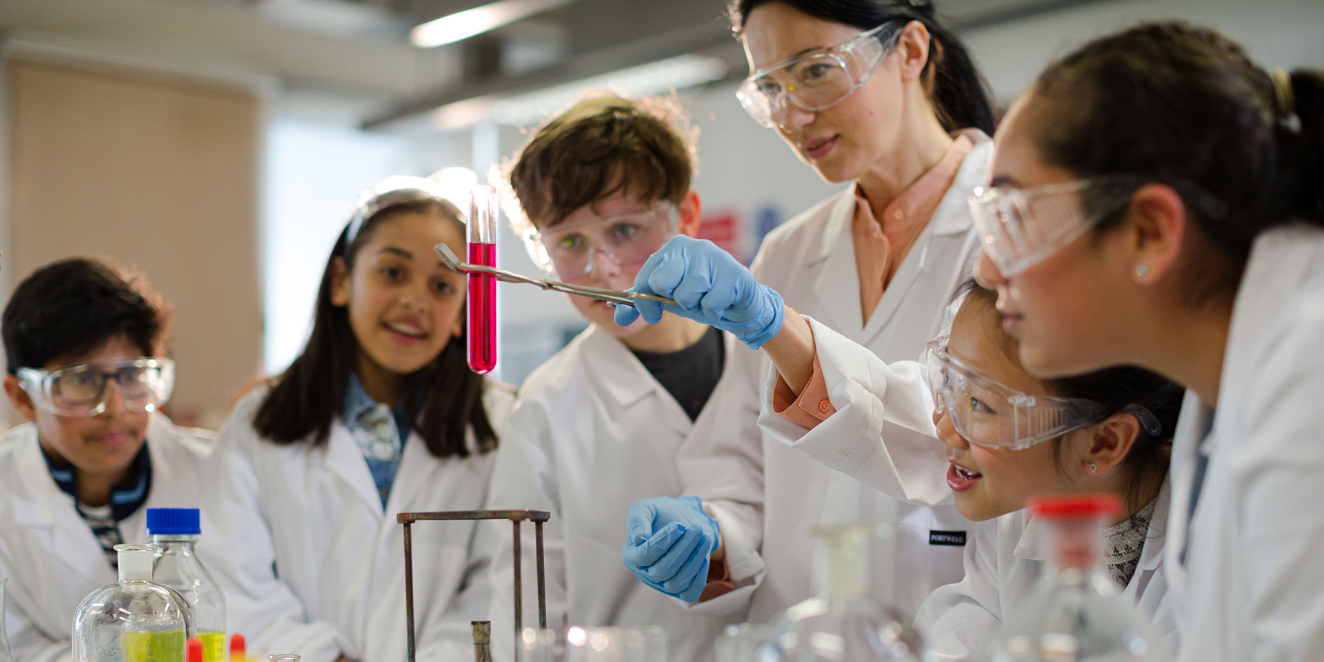 A female teacher and a group of students all in white lab coats and goggles conduct a science experiment.