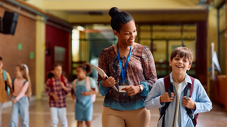 A black female teacher walks next to a young student in a school hallway