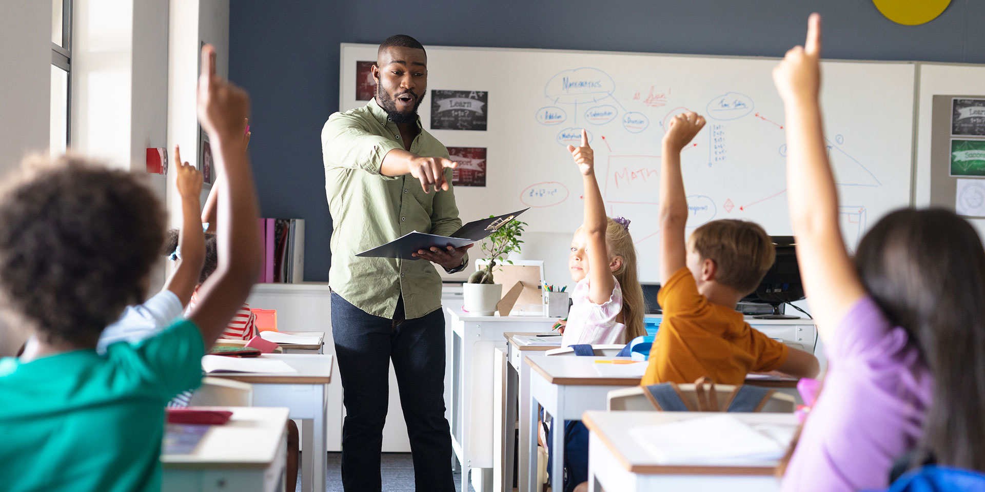 A black male teacher stands in front of a classroom pointing to a student with their hand raised