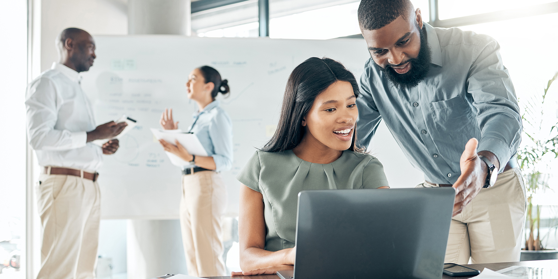 Students work on a laptop in the foreground, while two other students stand near a whiteboard in the background