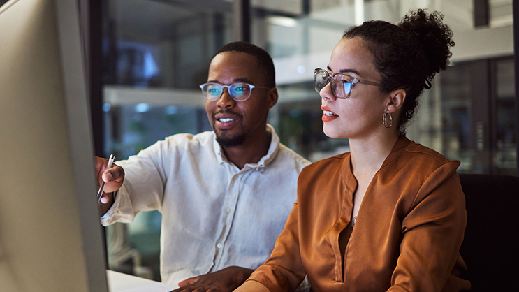 Two teachers working together in front of a computer. The male teacher is wearing a white button up and glasses while pointing at the computer screen. The female teacher is wearing glasses and an orange blouse while typing on a keyboard.