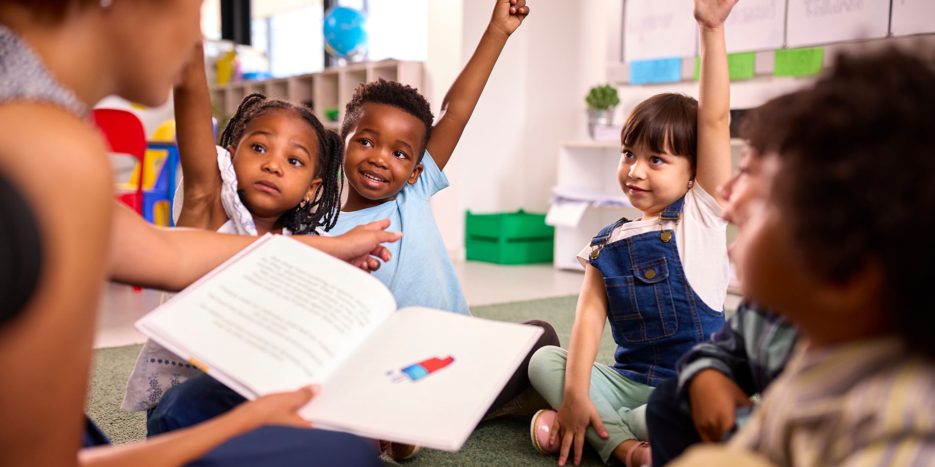 Students raise their hands while a teacher reads them a book