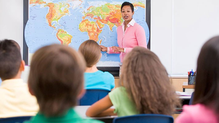 A female teacher stands at the front of a classroom pointing to a map