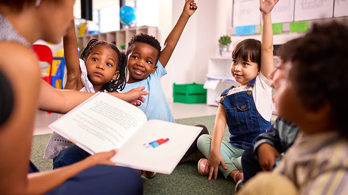 A teacher reads to a group of young students. Three have their hands raised