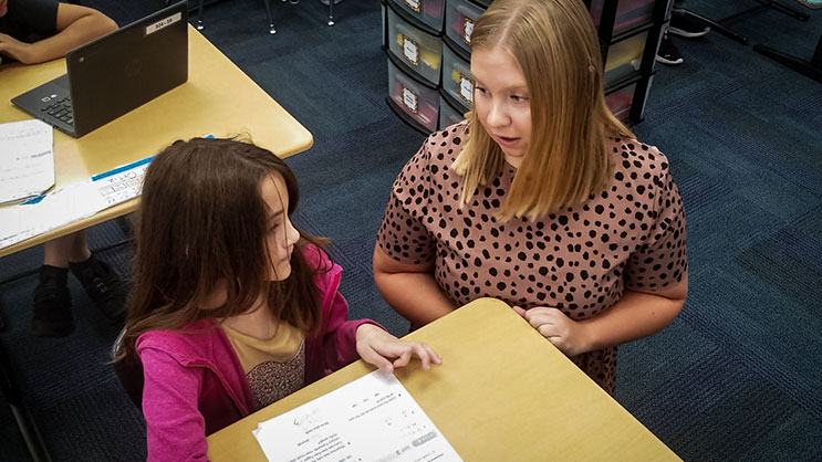 An undergraduate education student talking to an elementary student in a classroom.