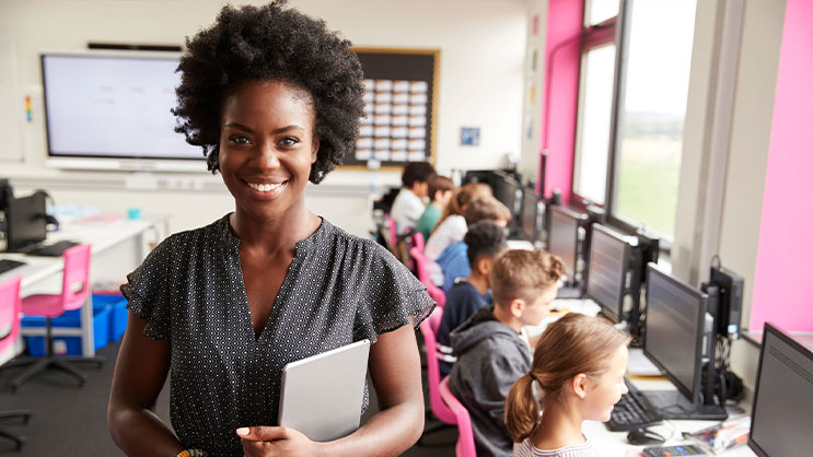 A school teacher facing the camera and wearing a black dress, multicolor bracelet, and holding a tablet. In the background are young students using computers.