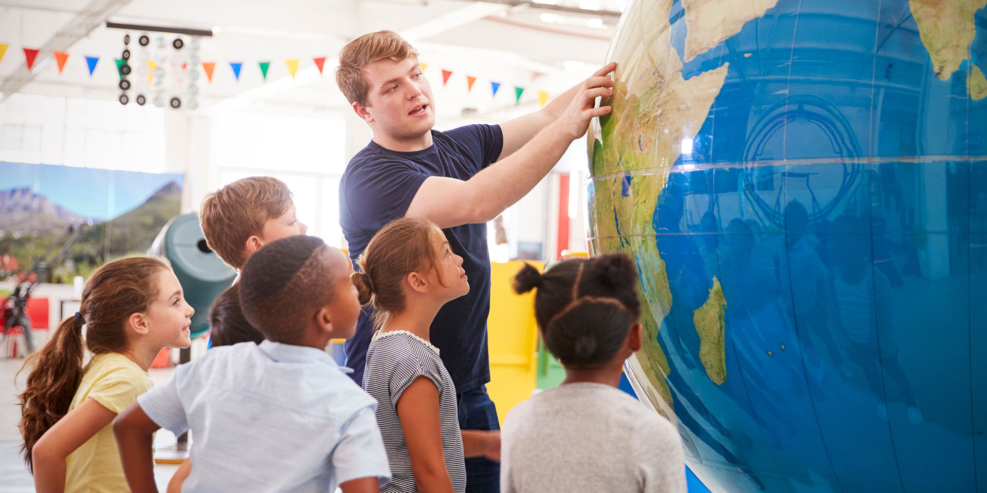 A male teacher surrounded by a group of students points to something on a very large globe