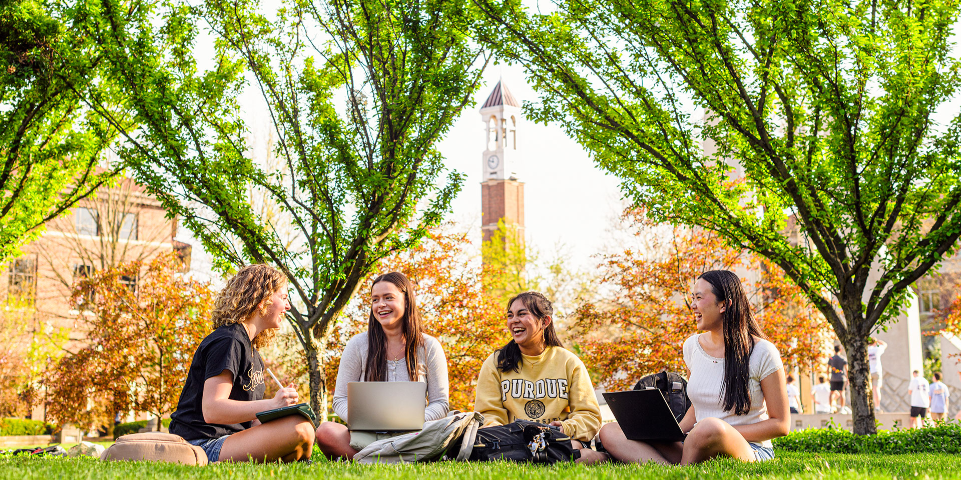 Four female Purdue University students sit outside on campus. Purdue's Bell Tower is seen in the distance behind them.