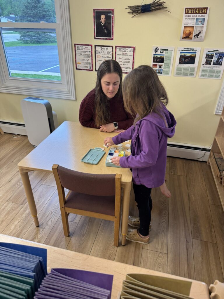 Teacher Candidate student Ali Drews seated at a table next to a young student. In front of them is a variety of finger paints.