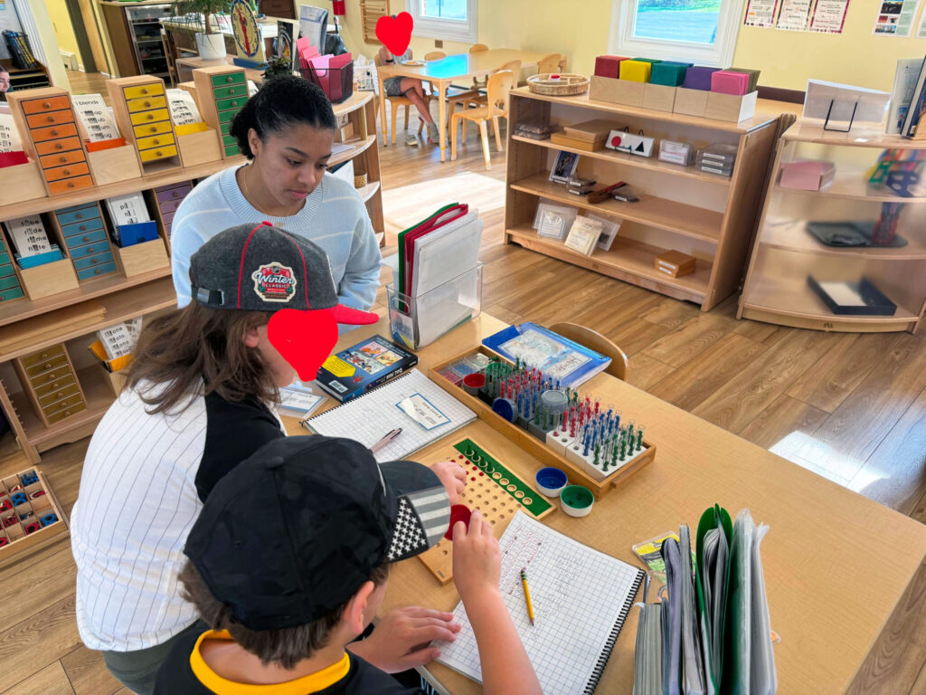 Teacher candidate stuent Jordan Garrison seated with two young students. Laying on a table in front of them is a game to practice arithmetic.